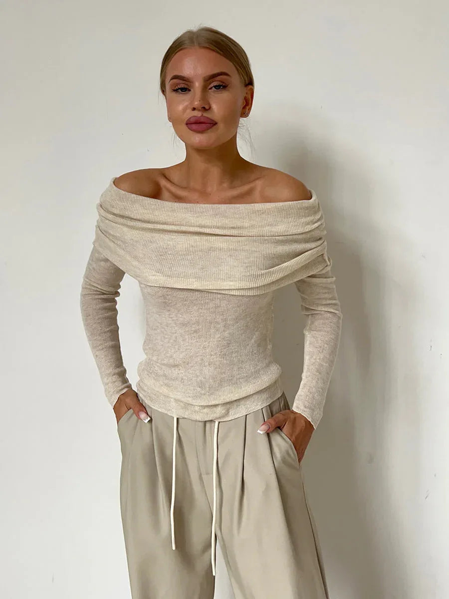 Woman wearing a cream off-the-shoulder ribbed knit sweater with long sleeves and beige wide-leg pants, hands in pockets, standing against a white wall.