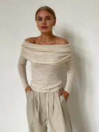Woman wearing a cream off-the-shoulder ribbed knit sweater with long sleeves and beige wide-leg pants, hands in pockets, standing against a white wall.
