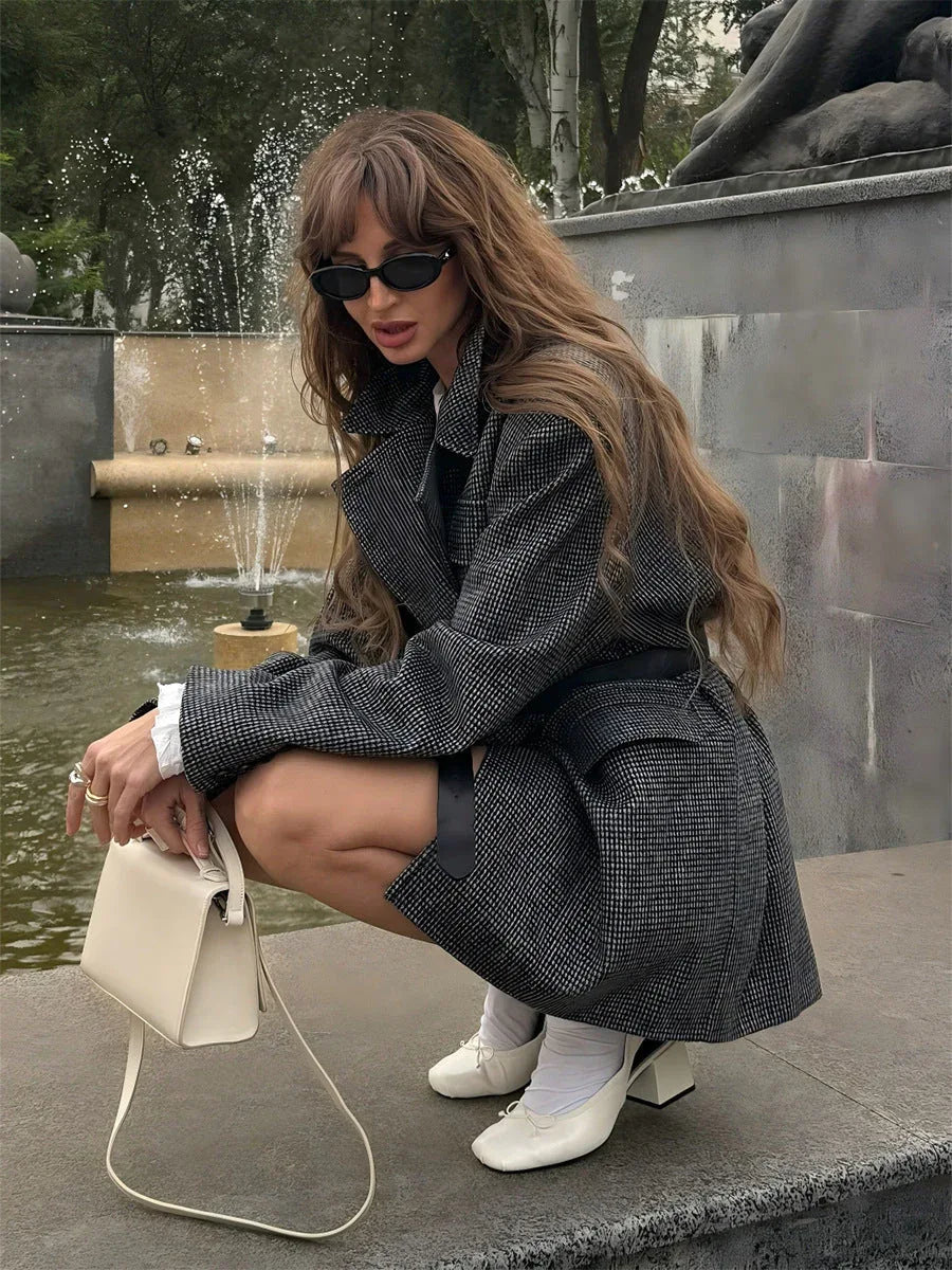 Woman crouching near a fountain wearing an oversized grey plaid double-breasted blazer, white t-shirt, shorts, white socks, and white block heels.