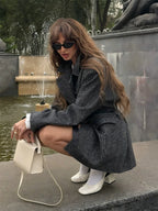 Woman crouching near a fountain wearing an oversized grey plaid double-breasted blazer, white t-shirt, shorts, white socks, and white block heels.