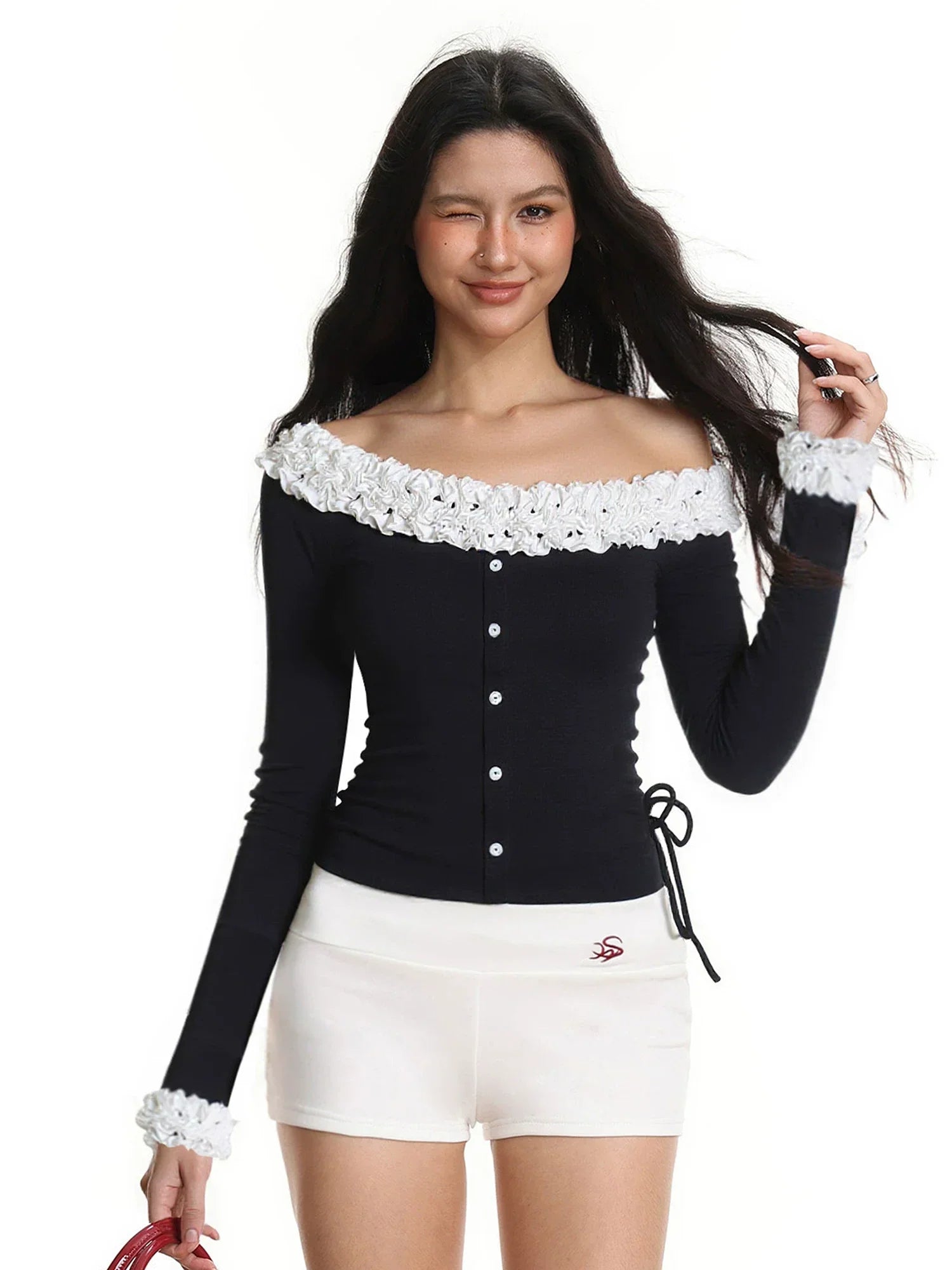 Model wearing a black off-the-shoulder long-sleeved top with white ruffled trim and white shorts, winking and touching her hair.