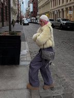 Person with pink hair walking on a cobblestone street, wearing a cream faux fur jacket and grey trousers.