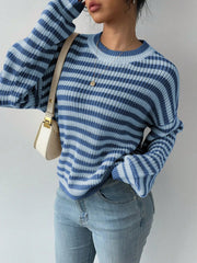 Woman wearing a blue and light blue striped knit sweater, light wash jeans, and a cream shoulder bag, with one hand on her hip and the other touching her hair.