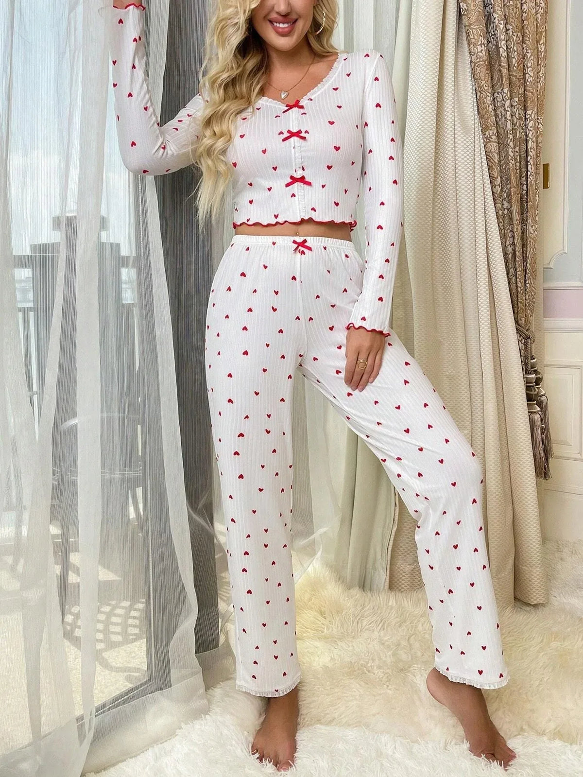 Woman wearing a white long-sleeve cropped pajama top and matching pants with a red heart print, standing by a window. Main image