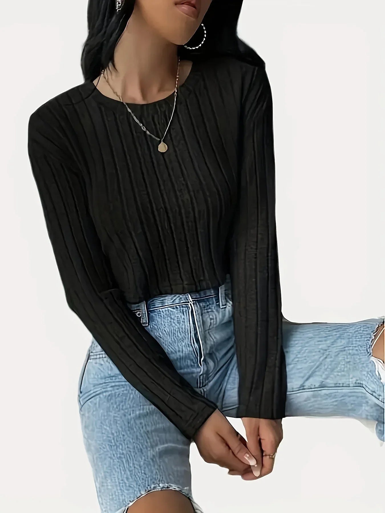 Woman wearing a black ribbed long-sleeve cropped top and light wash high-waisted jeans, sitting down with hands clasped in her lap.