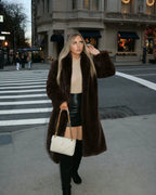 Woman standing at a crosswalk, wearing a long brown faux fur coat, beige turtleneck, and black faux leather mini skirt, holding a white quilted handbag.