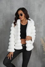 Woman wearing a white faux fur hooded jacket over a black crop top and dark jeans, with sunglasses on.