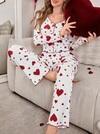 Woman wearing a white long-sleeved pajama top and pants with a red heart print, sitting on a couch and holding a red heart pillow.