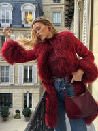 Woman standing on a balcony, wearing a red suede coat with fluffy red fur trim and cuffs, a red turtleneck, and blue jeans, holding a red handbag.
