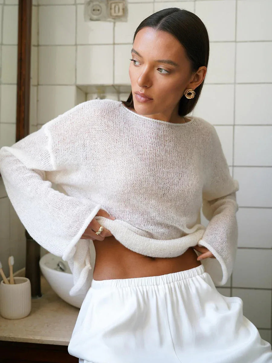 Woman wearing a white sheer knit sweater and white pants, lifting the sweater to reveal her midriff.