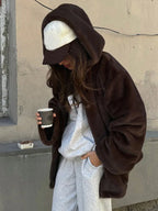 Woman wearing a brown faux fur hooded jacket over a grey hoodie and sweatpants, with a white baseball cap, holding a coffee cup.