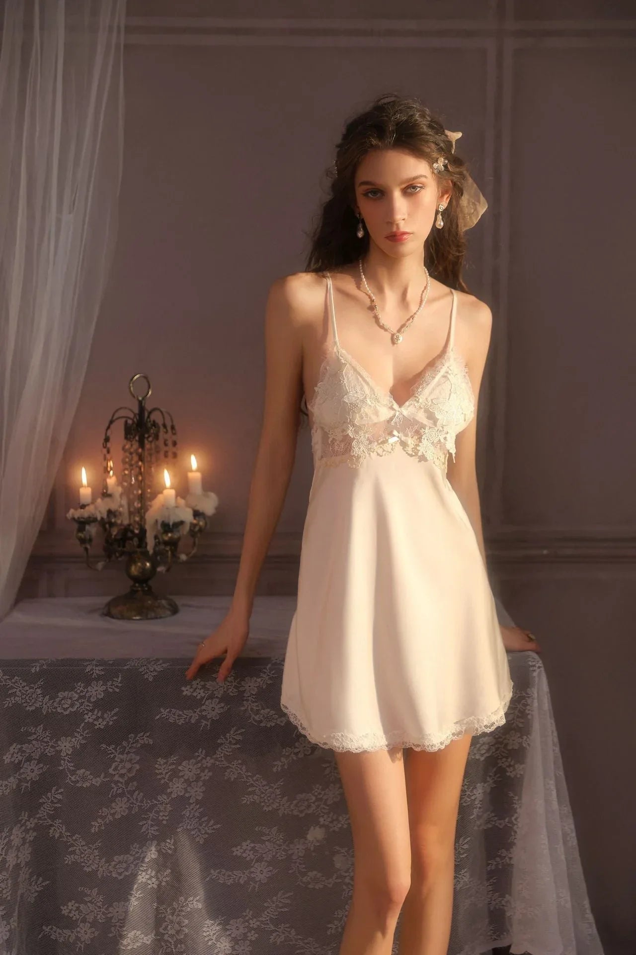 Woman wearing a white satin babydoll nightgown with lace trim and a matching robe, standing next to a table with candles.
