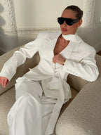 Woman sitting in a chair wearing a white oversized blazer and trousers, accessorized with sunglasses.