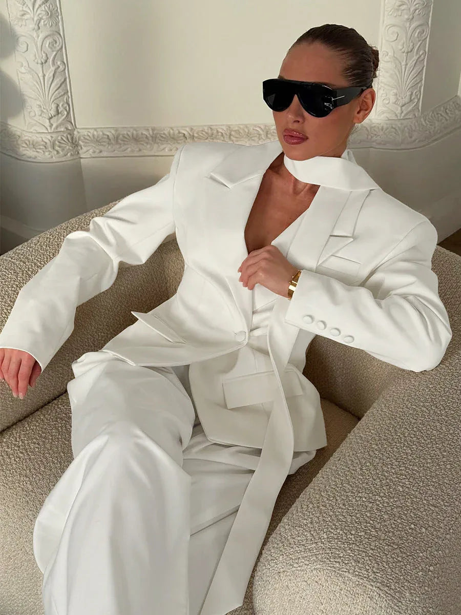 Woman sitting in a chair wearing a white oversized blazer and trousers, accessorized with sunglasses.