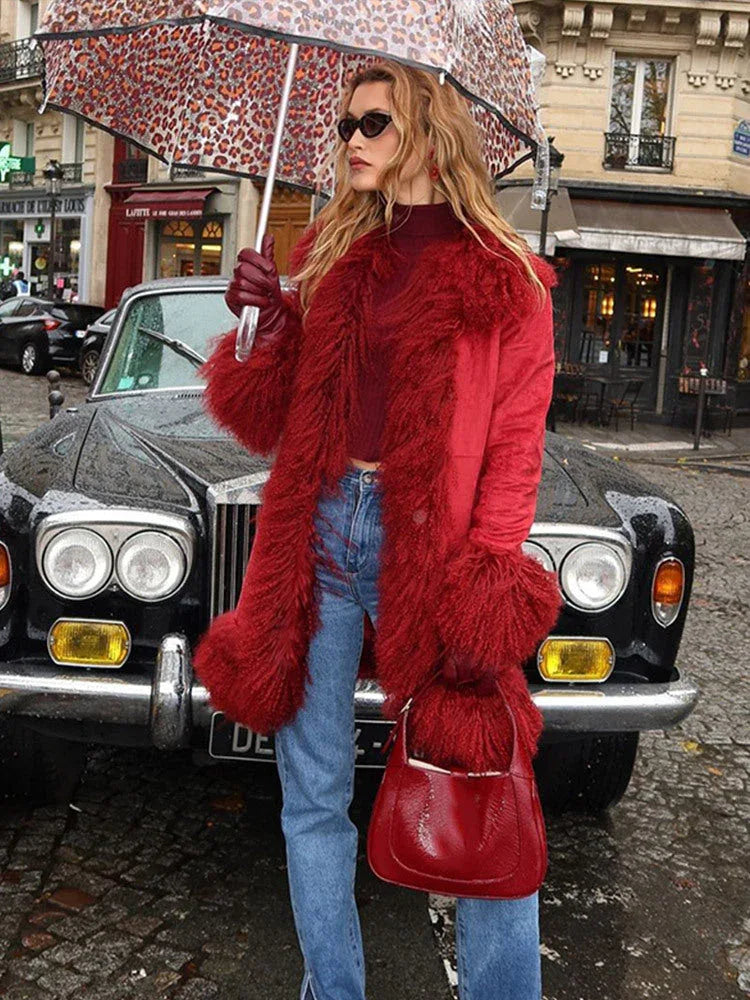 Woman standing in front of a vintage car, holding a leopard print umbrella, wearing a red suede coat with fluffy red fur trim and cuffs, a red turtleneck, blue jeans, and red gloves.