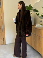 Woman in a brown faux fur hooded jacket and brown wide-leg pants, holding a coffee cup indoors.