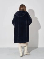 Woman wearing a black hooded faux fur coat, cream dress, and white boots, back view.