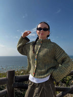 Woman wearing a green and black striped knit cardigan with colorful buttons, posing with hand raised, by the ocean.