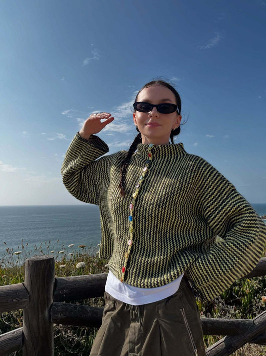 Woman wearing a green and black striped knit cardigan with colorful buttons, posing with hand raised, by the ocean.