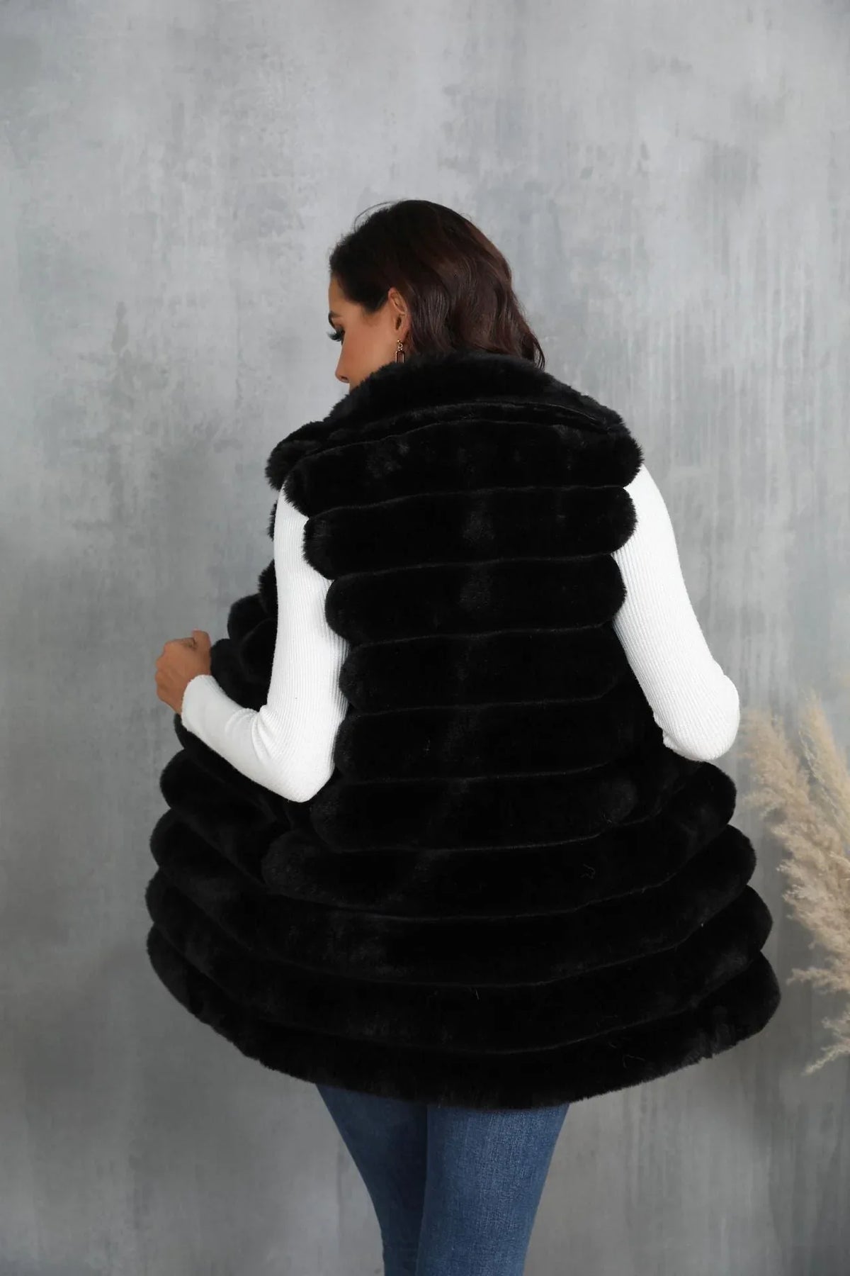 Back view of a woman wearing a black faux fur vest over a white turtleneck and blue jeans.