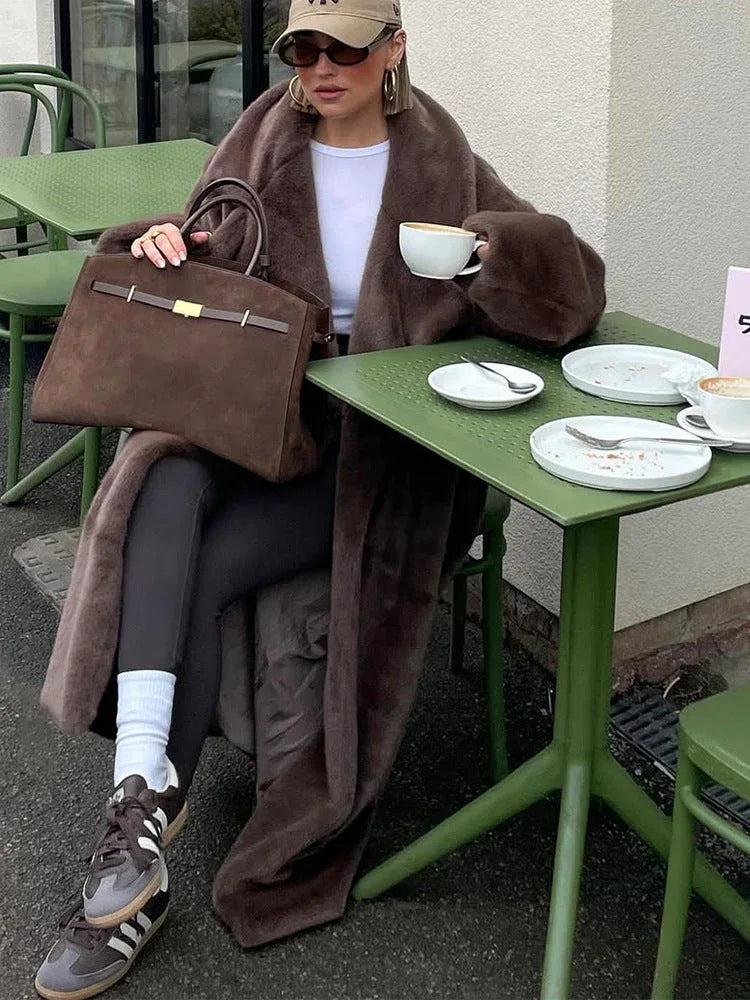 A close-up of a woman sitting at an outdoor cafe table, holding a coffee cup and wearing a brown faux fur coat.