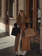 Woman in a camel coat with faux fur trim, holding two shopping bags and a tan handbag.