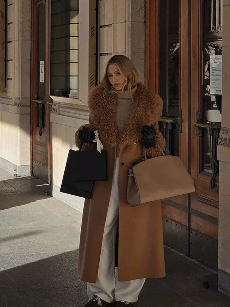 Woman in a camel coat with faux fur trim, holding two shopping bags and a tan handbag.