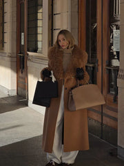 Woman in a camel coat with faux fur trim, holding two shopping bags and a tan handbag.