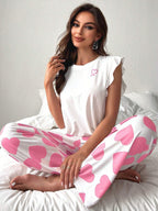 Woman sitting on a bed wearing white pajamas with pink heart print pants and a white top with a pink ribbon graphic.