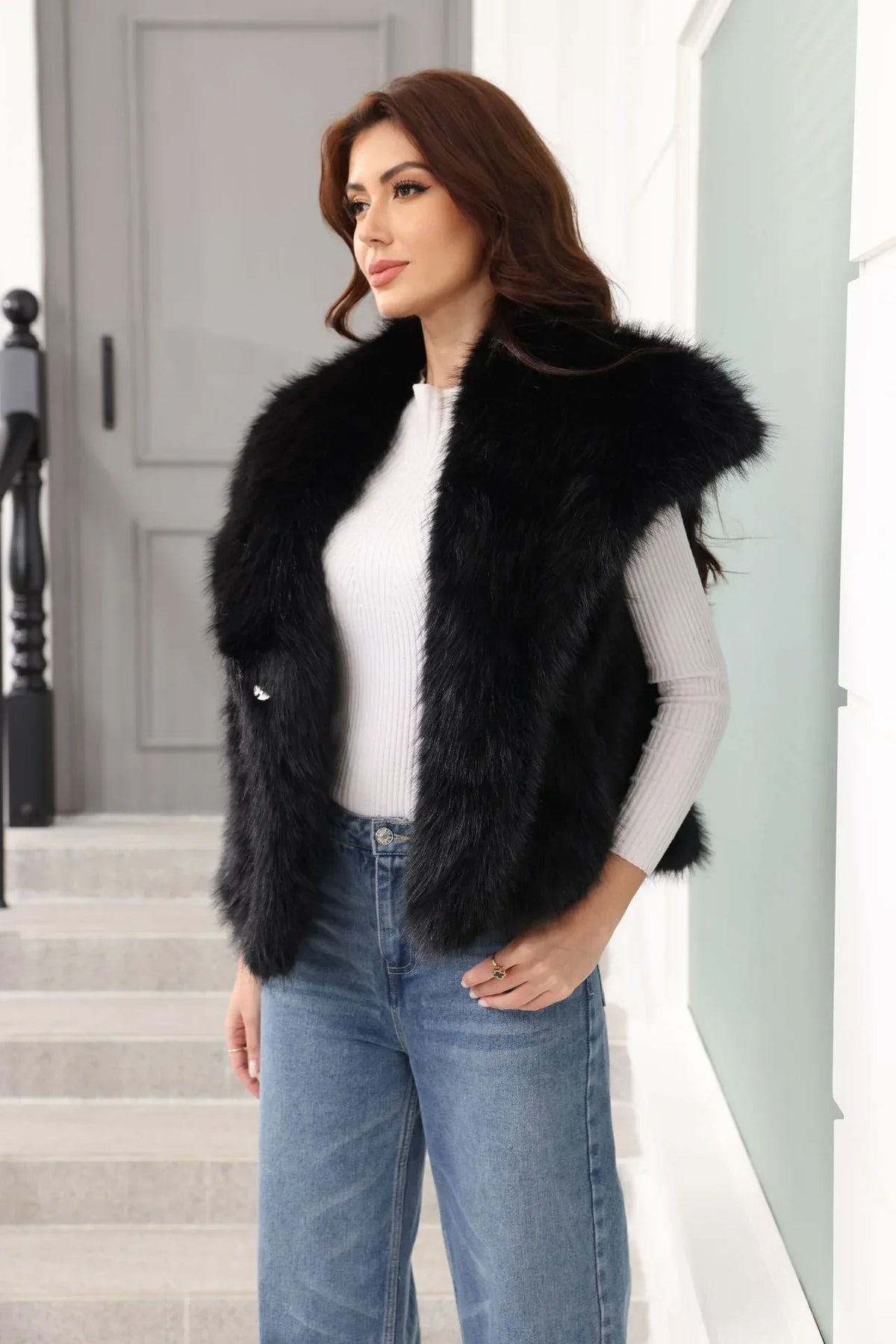 Woman wearing a black faux fur vest over a white long-sleeved shirt and blue jeans, with hands in pockets
