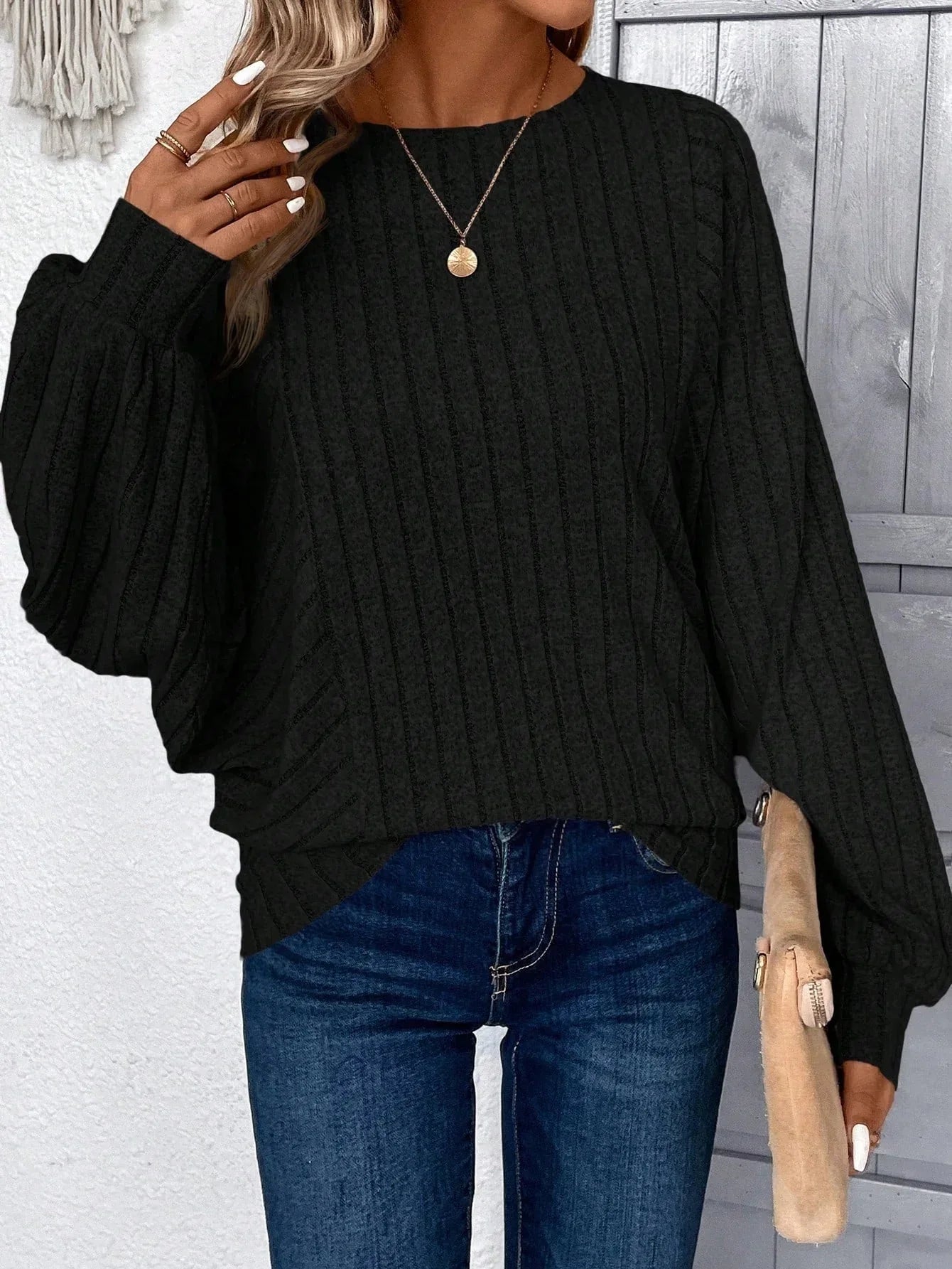Woman in a black ribbed knit sweater, blue jeans, and a gold pendant necklace, posing with her hand near her face.