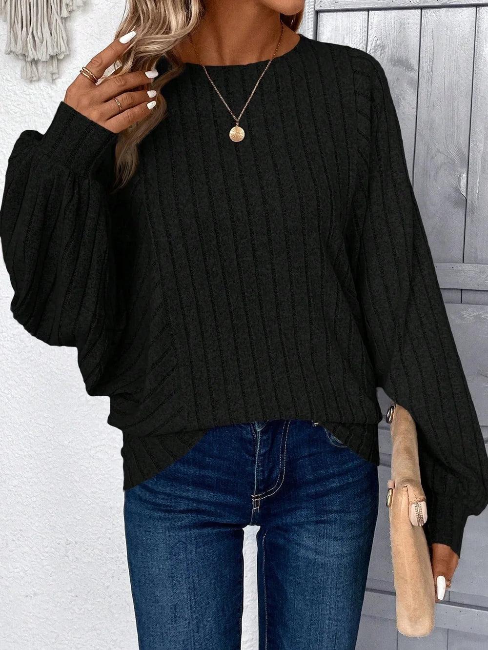 Woman in a black ribbed knit sweater, blue jeans, and a gold pendant necklace, posing with her hand near her face.