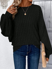 Woman in a black ribbed knit sweater, blue jeans, and a gold pendant necklace, posing with her hand near her face.