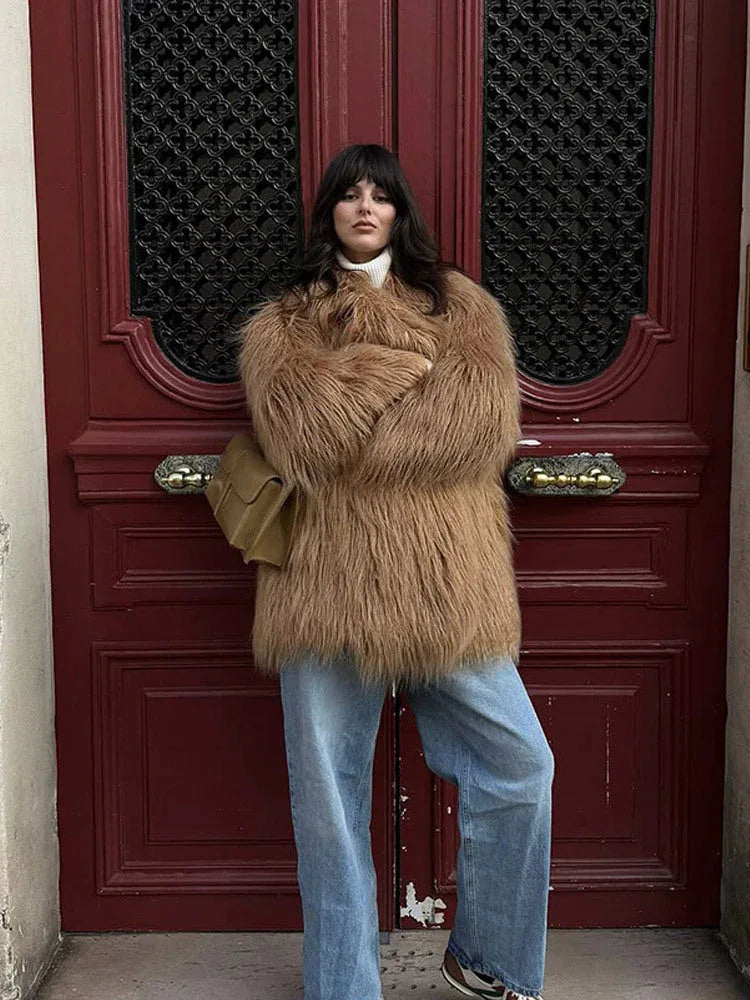 woman wearing a shaggy brown faux fur coat and blue jeans, standing in front of a red door