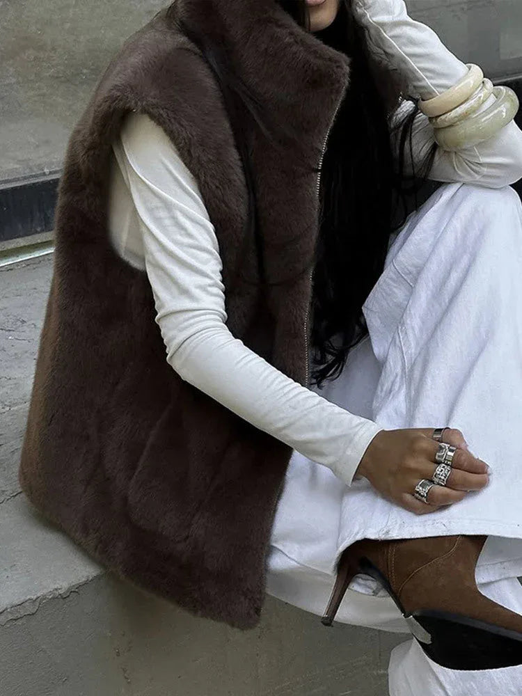 Woman in a brown faux fur vest, white long-sleeved shirt, and white pants sitting on steps.