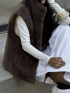 Woman in a brown faux fur vest, white long-sleeved shirt, and white pants sitting on steps.