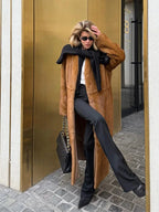 Woman in a long brown faux fur coat, white shirt, black pants, and black scarf, holding a quilted black handbag.
