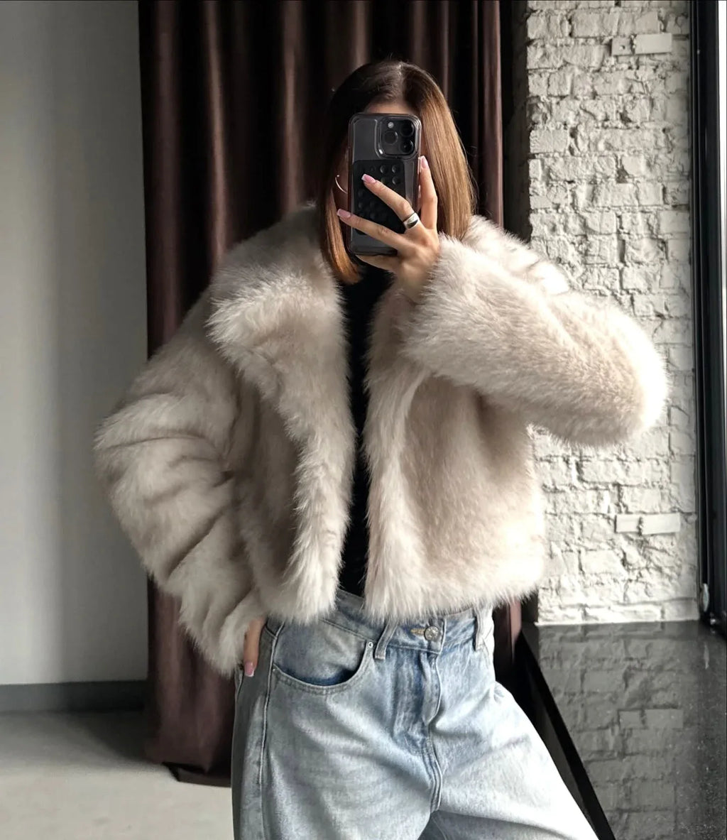 Close-up of a woman wearing a cream faux fur cropped jacket and light wash wide-leg jeans, holding a phone to take a mirror selfie.