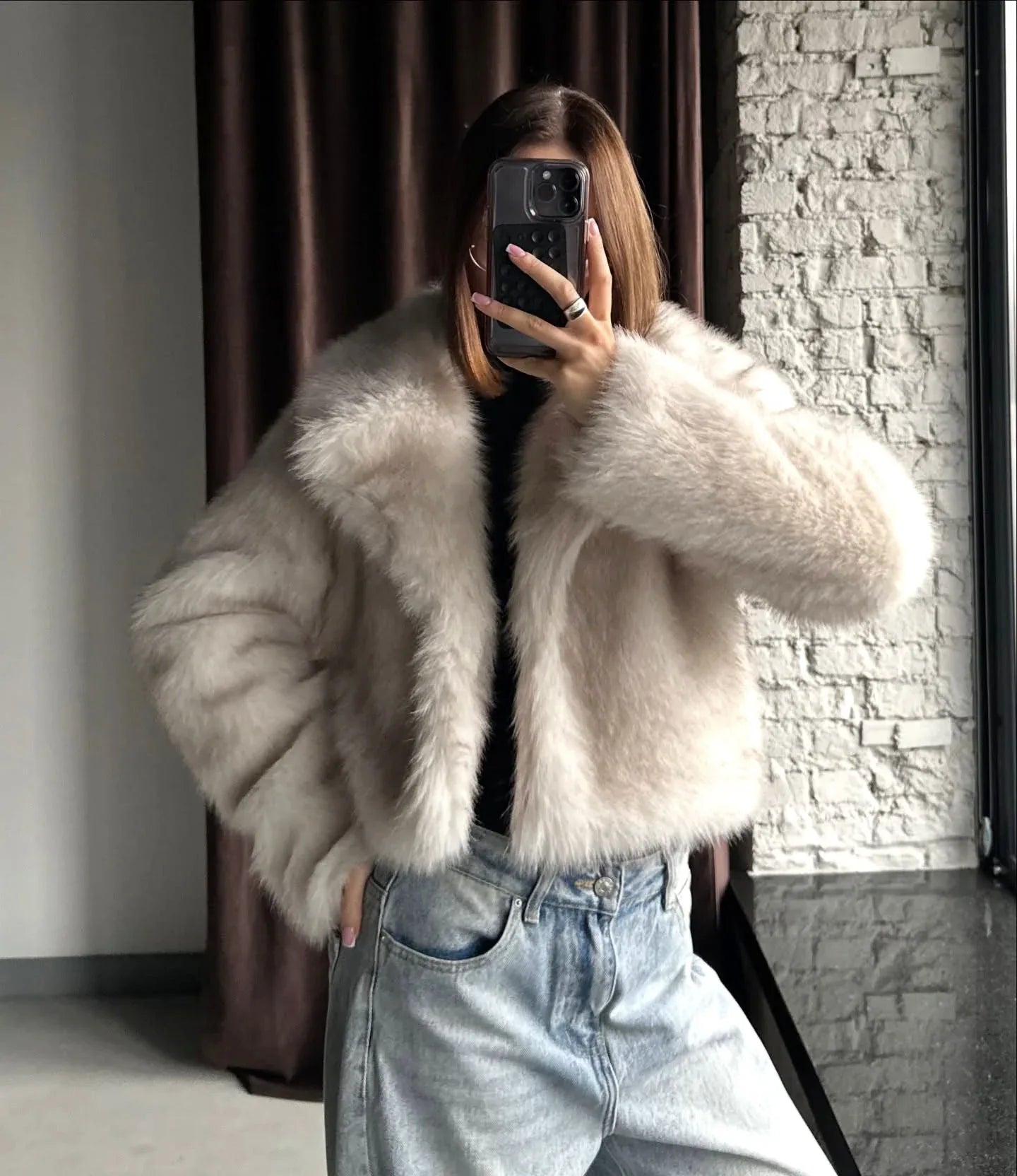 Close-up of a woman wearing a cream faux fur cropped jacket and light wash wide-leg jeans, holding a phone to take a mirror selfie.
