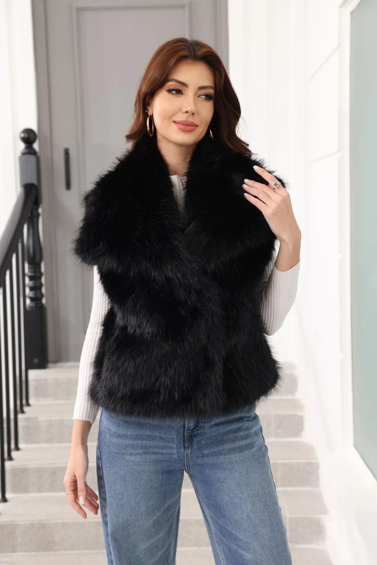 Woman wearing a black faux fur vest over a white long-sleeved shirt and blue jeans, standing on stairs