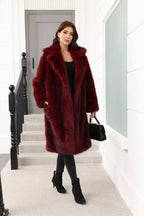 A woman wearing a long, burgundy faux fur coat over a black top and black pants, holding a black handbag.