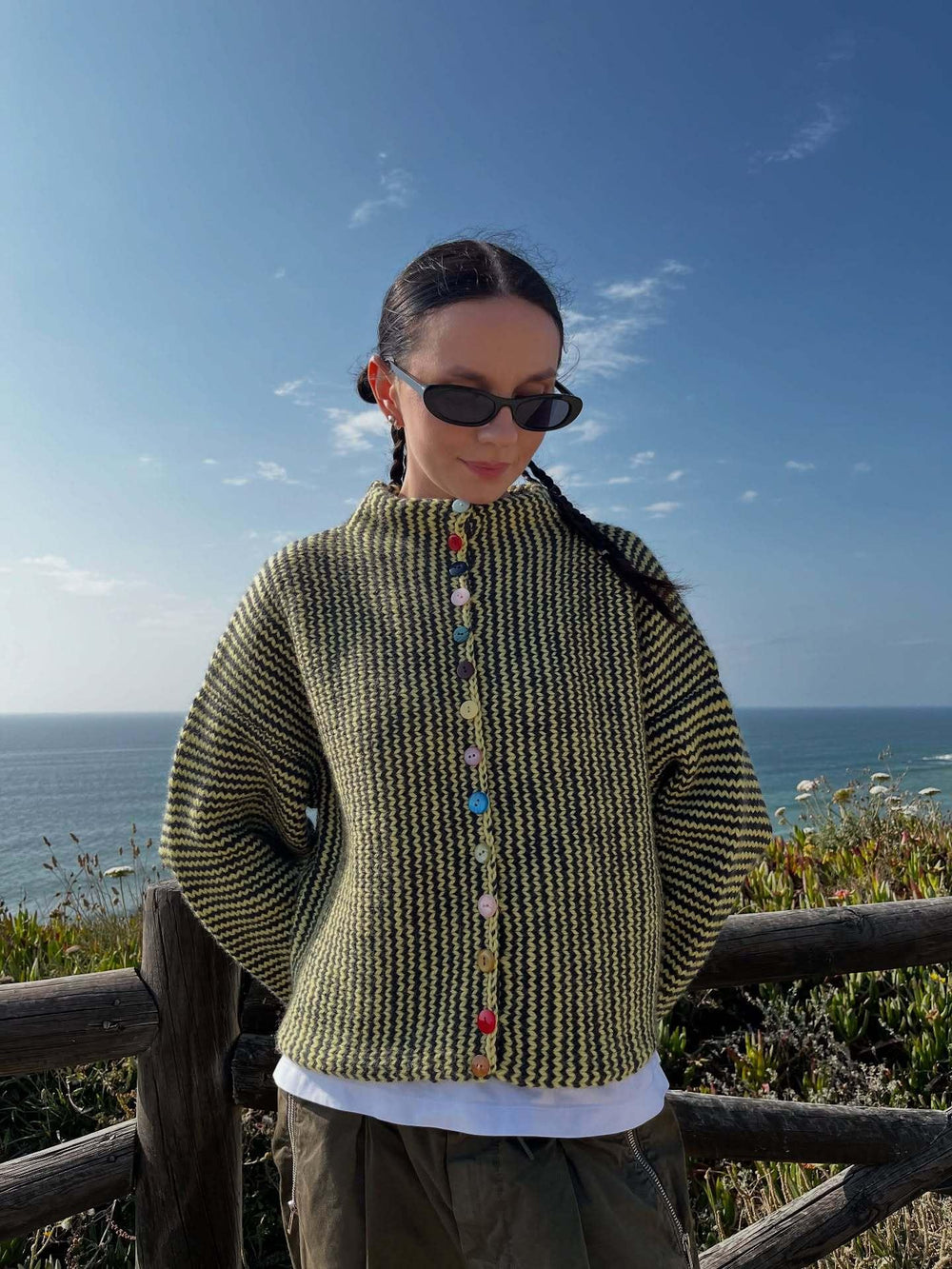 Woman wearing a green and black striped knit cardigan with colorful buttons, standing by the ocean.