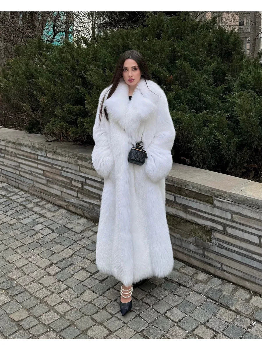 Woman wearing a long white faux fur coat with a large collar and a black quilted handbag, standing on a cobblestone path. Main image