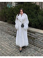 Woman wearing a long white faux fur coat with a large collar and a black quilted handbag, standing on a cobblestone path.
