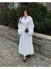 Woman wearing a long white faux fur coat with a large collar and a black quilted handbag, standing on a cobblestone path.