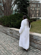 Rear view of a woman in a long white faux fur coat walking along a stone wall and green bushes.