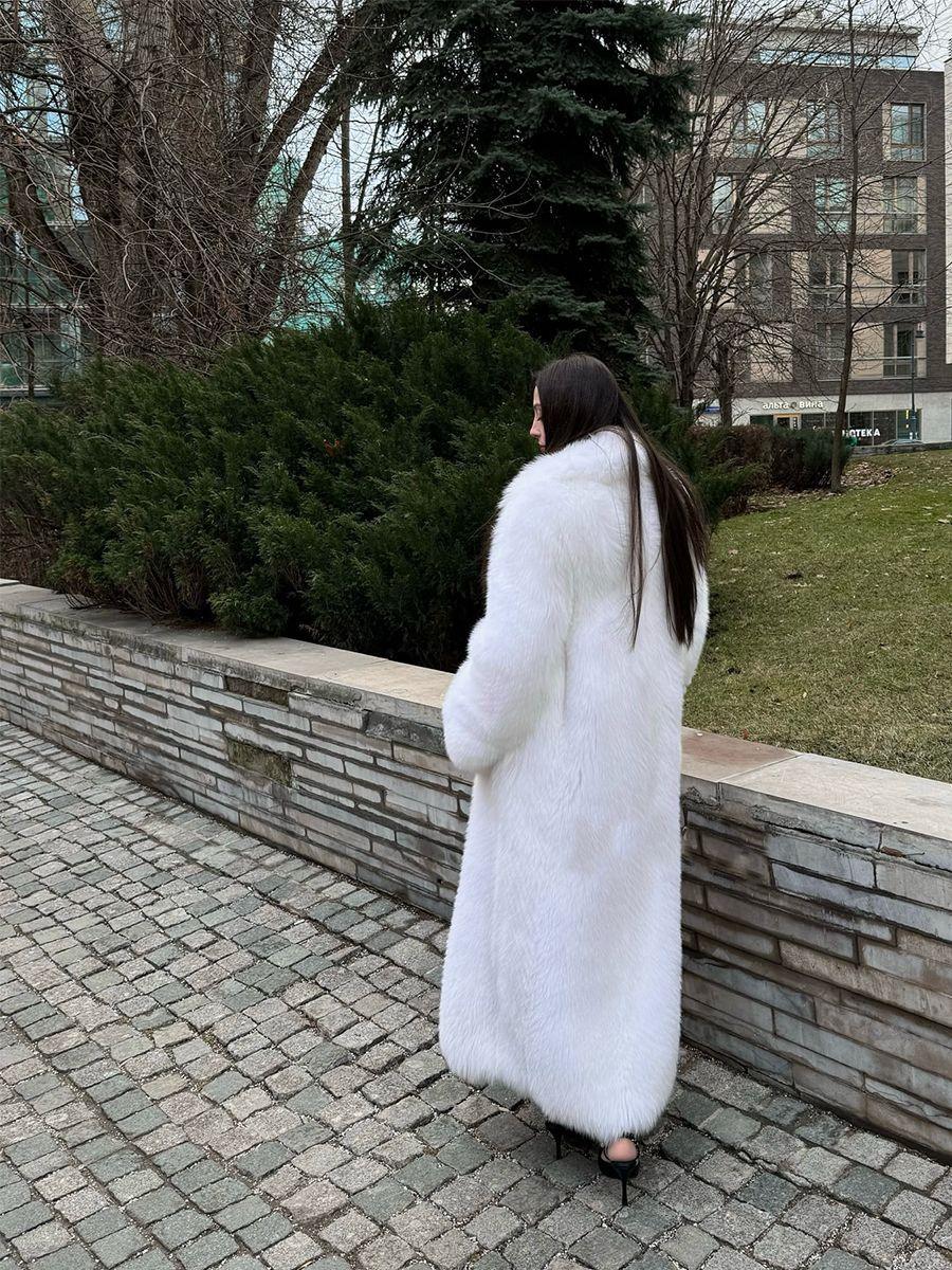 Rear view of a woman in a long white faux fur coat walking along a stone wall and green bushes.