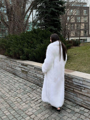 Rear view of a woman in a long white faux fur coat walking along a stone wall and green bushes.
