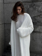 Woman wearing a long white fuzzy knit cardigan and a striped dress, standing against a stone wall.