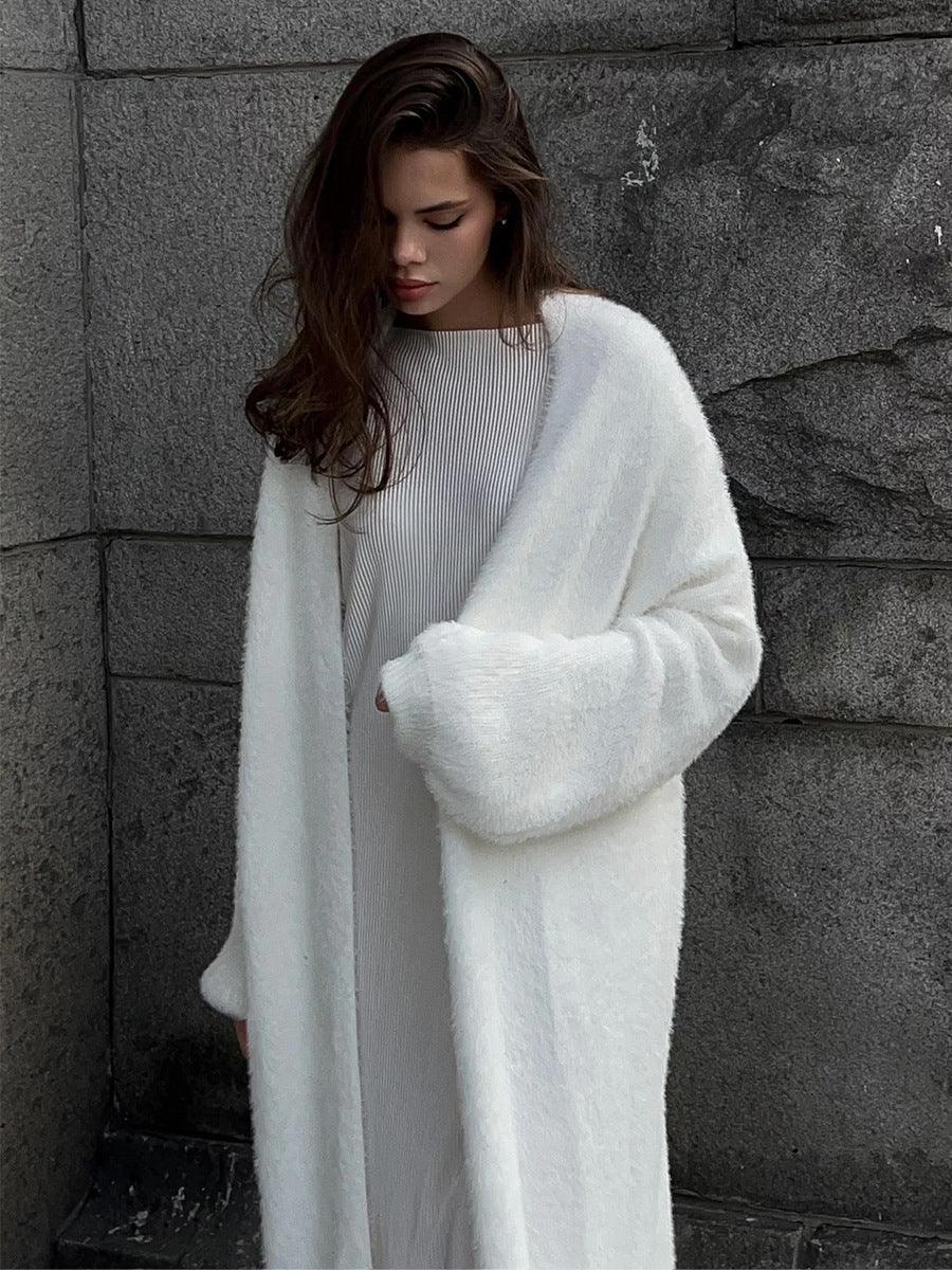 Woman wearing a long white fuzzy knit cardigan and a striped dress, standing against a stone wall.